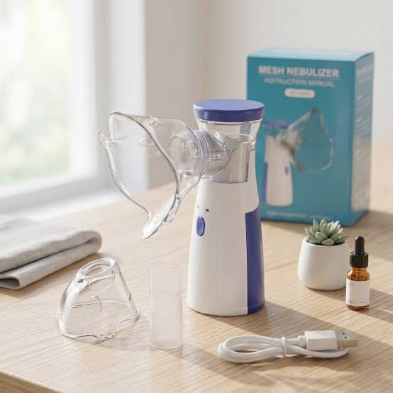 A handheld mesh nebulizer set displayed on a light wood table near a window, including the main device, clear face mask, tubing, small accessory cup, USB charging cable, folded towel, a small dropper bottle, a potted succulent, and the product box.