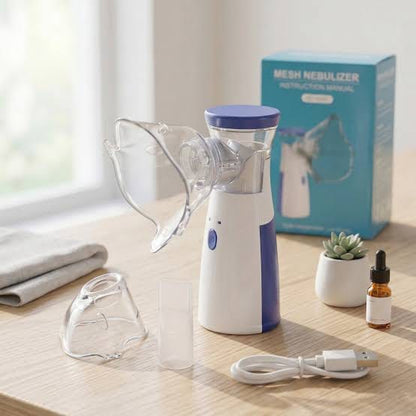 A handheld mesh nebulizer set displayed on a light wood table near a window, including the main device, clear face mask, tubing, small accessory cup, USB charging cable, folded towel, a small dropper bottle, a potted succulent, and the product box.